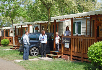 Family stands by SunLodge Maple at Camping Bella Italia, Italy, with a car and trees outside wooden lodges.