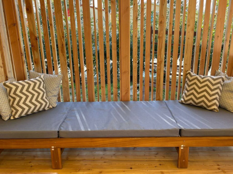 Outdoor bench with gray cushions and patterned pillows at SunLodge Babul, surrounded by wooden slats and greenery.