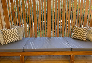 Banco al aire libre con cojines grises y almohadas decorativas en SunLodge Babul, rodeado de listones de madera y vegetación.