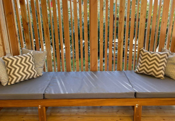 Banco al aire libre con cojines grises y almohadas decorativas en SunLodge Babul, rodeado de listones de madera y vegetación.