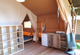 Interior de una tienda safari con cocina, estantería y cama en Camping Oos Heem, Bélgica.