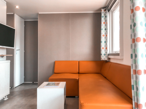 Hedendaagse woonkamer in Forest lodge bij Vakantiepark De Luttenberg met oranje zetel en TV.