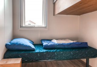 Eenvoudig bed met blauw bedlinnen in het Forest lodge van Vakantiepark De Luttenberg, Nederland, bij een raam.