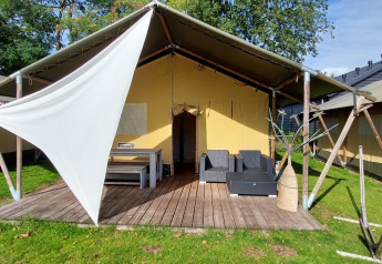 Safari telt lodge med træterrasse, havemøbler og skyggeelement, omgivet af grønne græsplæner og træer.
