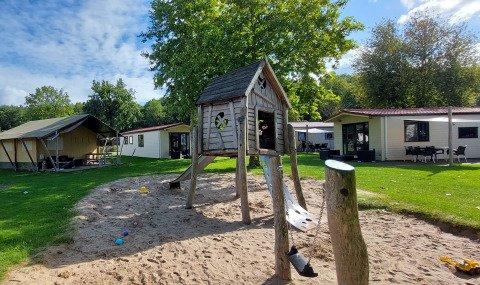 Parque infantil con casa de madera y arena junto a una tienda safari en Vakantiepark De Luttenberg, Países Bajos.