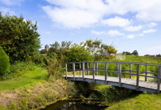 Wooden bridge over a small stream at Camping Waddenzee, with lush greenery and a blue sky in North Holland.