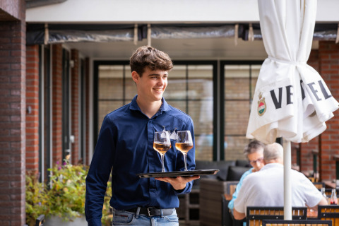 Joven camarero con camisa azul sirviendo dos copas de vino en la terraza de Camping Waddenzee, Holanda Septentrional.