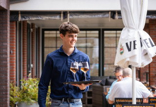Giovane cameriere con camicia blu serve due bicchieri di vino sulla terrazza del Camping Waddenzee, Olanda Nord.