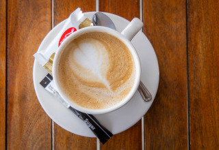 En kop cappuccino med latte art, sukkerpakker og en kiks på en træbord ved Camping Waddenzee, Holland.