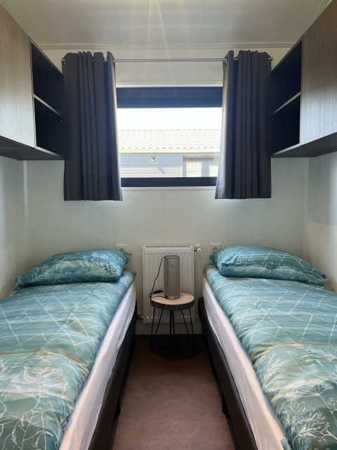 Dos camas individuales con ropa de cama verde, ventana con cortinas y mesa de noche en la cabaña De Parel.