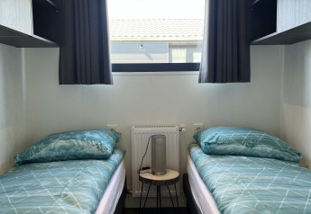 Dos camas individuales con ropa de cama verde, ventana con cortinas y mesa de noche en la cabaña De Parel.