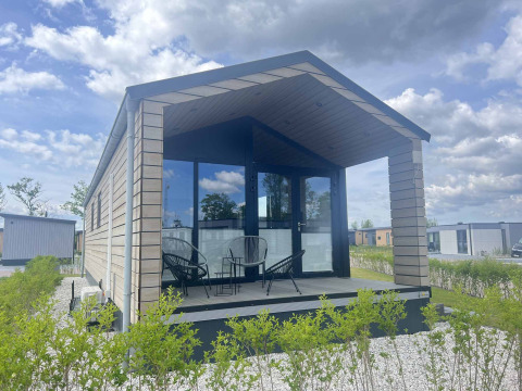 Lodge moderne à De Parel avec façade vitrée, terrasse couverte et sièges extérieurs sous un ciel bleu.