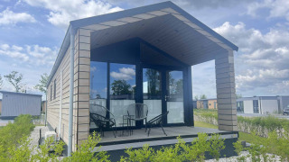 A modern lodge at De Parel featuring a glass front, covered terrace, and outdoor seating under a blue sky.