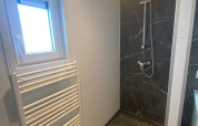 Baño moderno con ducha, ventana, radiador toallero y azulejos oscuros en una cabaña en los Países Bajos.