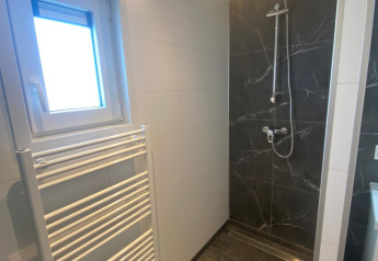 Baño moderno con ducha, ventana, radiador toallero y azulejos oscuros en una cabaña en los Países Bajos.