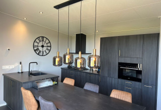 Moderne keuken met donkere kasten, eettafel, hanglampen en wandklok in lodge De Zeester, Nederland.