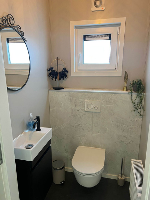 Baño moderno con inodoro suspendido, lavabo pequeño, espejo redondo y ventana en De Zeester, Países Bajos.