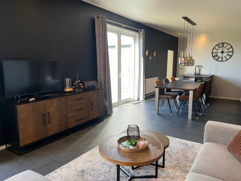 Modern lodge living and dining area with black wall, TV, dining table, and large windows at De Zeester, Netherlands.