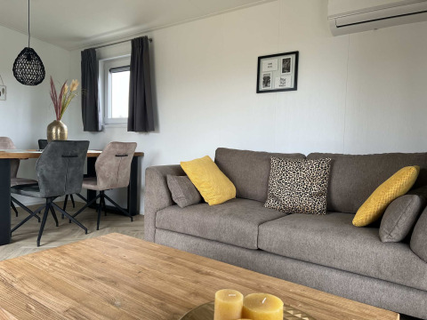 Modernes Wohnzimmer in der Dijkzicht-Lodge mit grauem Sofa, gelben Kissen, Esstisch und großen Fenstern.