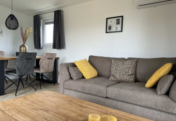 Modern living room at Dijkzicht lodge with a gray sofa, yellow cushions, dining table, and large windows.
