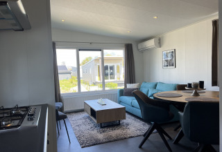 Moderne stue i Lodge Noordzee med blå sofa, spisebord og store vinduer, Vakantiepark de Zeeuwse Parel.