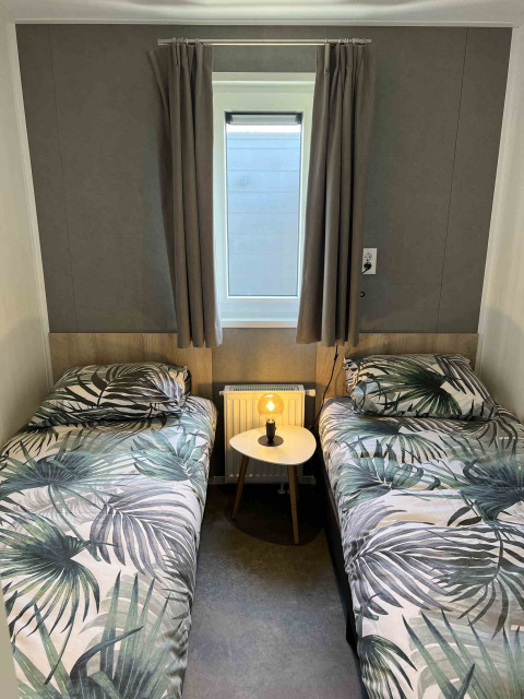 Dos camas individuales con ropa de cama tropical, mesa pequeña con lámpara y ventana en una cabaña moderna.