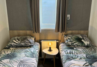 Dos camas individuales con ropa de cama tropical, mesa pequeña con lámpara y ventana en una cabaña moderna.