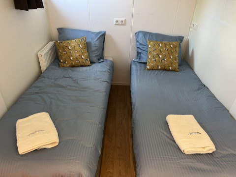 Dos camas individuales con sábanas azules, cojines amarillos y toallas en Parkzicht, Vakantiepark de Zeeuwse Parel.