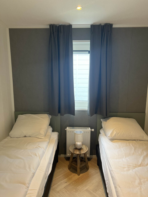 Dos camas individuales con sábanas blancas, mesa pequeña con lámpara y ventana cubierta con cortinas oscuras.