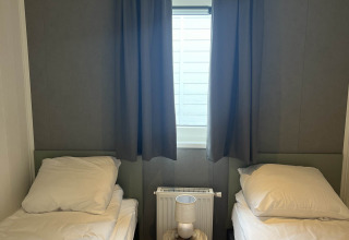 Dos camas individuales con sábanas blancas, mesa pequeña con lámpara y ventana cubierta con cortinas oscuras.