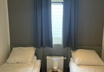 Deux lits simples avec draps blancs, une petite table avec lampe et une fenêtre centrale avec rideaux sombres.