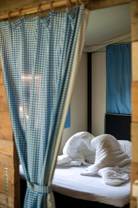 Vista de una cama con ropa de cama blanca desordenada en una tienda safari con cortinas a cuadros azules.