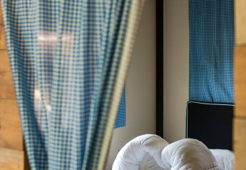 View of a bed with messy white bedding in a safari tent, blue checkered curtains and windows.