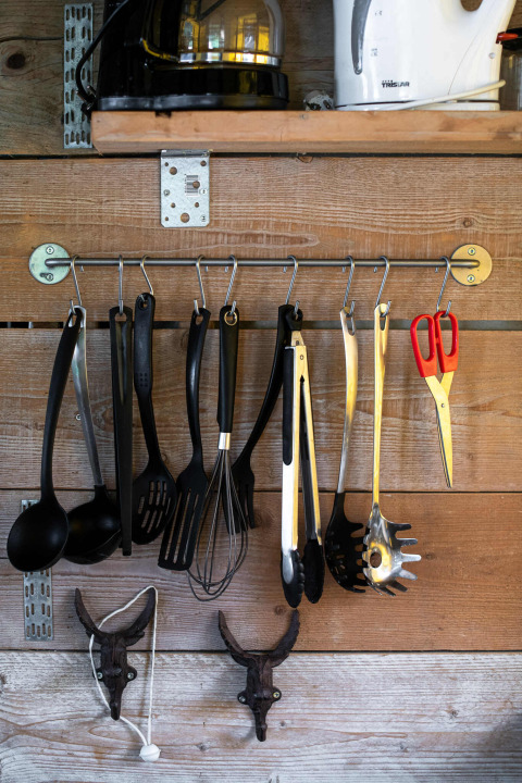 Ustensiles de cuisine accrochés à des crochets sur un mur en bois sous une étagère dans une tente safari Tentlodge.