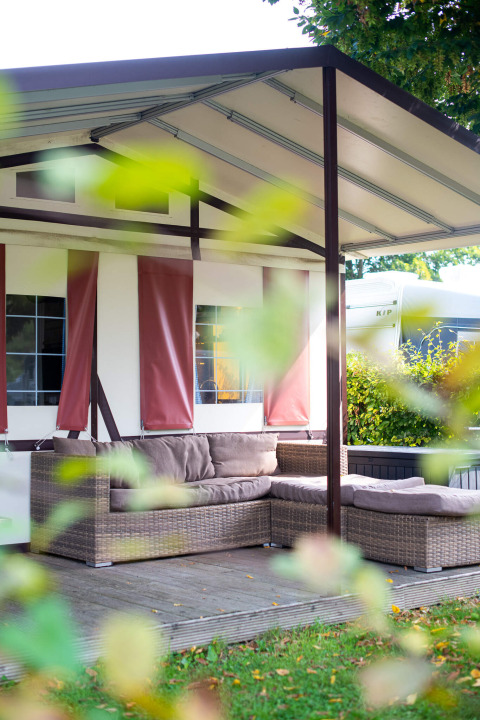Udendørs siddeplads med rattan-sofa foran en safari-tentlodge med lyse gardiner og grønne planter.