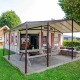 Safari-teltet Tentlodge med overdækket terrasse, træbord og loungemøbler på en grøn campingplads.