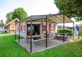 Safari-Zelt Tentlodge mit überdachter Terrasse, Holztisch und Lounge-Möbeln auf grünem Campingplatz.