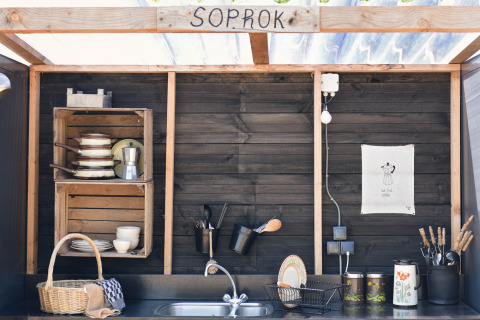 Outdoor wooden kitchen area with black wall, shelves, dishes, and utensils at Smûk Lytse Bell Tent.