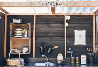 Udendørs køkkenområde i træ med sort væg, hylder, service og redskaber ved Smûk Lytse Bell Tent.