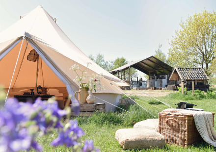 Smûk Lytse Bell Tent glamping tafereel: open tent met kussens, mand en boerderij op de achtergrond.