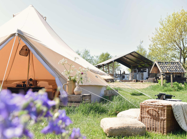 Smûk Lytse Bell Tent glamping tafereel: open tent met kussens, mand en boerderij op de achtergrond.