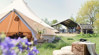 Scène de glamping à la Smûk Lytse Bell Tent avec tente, paniers, coussins et bâtiments de ferme en fond.