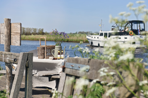 Table de pique-nique extérieure au bord de l’eau avec fleurs, boissons et bateau passant en arrière-plan.