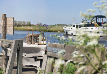 Picknicktafel aan het water met bloemen en drank, boot op de achtergrond op een zonnige dag in Nederland.