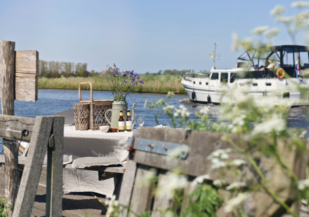 Picknicktafel aan het water met bloemen en drankjes, boot vaart voorbij op de achtergrond op zonnige dag.