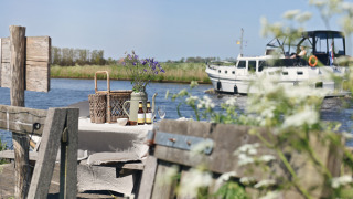 Table de pique-nique extérieure au bord de l’eau avec fleurs, boissons et bateau passant en arrière-plan.