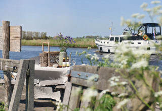 Outdoor picnic table by the water with drinks and flowers, boat passing in the background on a sunny day.