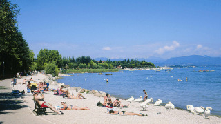 Playa soleada cerca de Peschiera del Garda, Italia, con personas tomando el sol y cisnes junto al lago.