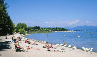 Playa soleada cerca de Peschiera del Garda, Italia, con personas tomando el sol y cisnes junto al lago.