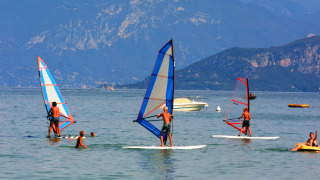 Turistas disfrutan de windsurf y natación en el lago con montañas al fondo en Camping Bella Italia, Véneto.